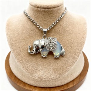 May include: Silver-toned elephant pendant necklace with a light blue opal body and decorative saddle. The elephant has detailed features and is suspended from a silver chain. The necklace is displayed on a beige bust.