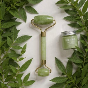 Op de afbeelding: Een jade gezichtsroller met goudkleurige metalen accenten en een klein potje groene crème met de tekst "Face Balm Healer" op een wit oppervlak, omringd door groene bladeren. De roller is ongeveer 15 cm lang.