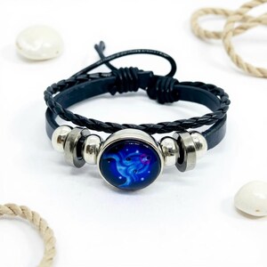 May include: Black leather bracelet with a braided band and a round, blue and purple design in the center. The bracelet has silver-toned metal accents and a black cord closure. The bracelet is on a white surface with decorative elements.