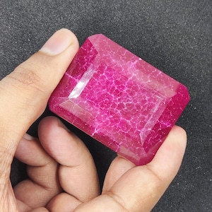 Könnte beinhalten: Ein großer, rechteckiger, facettierter Edelstein in tiefem Magenta. Der Edelstein weist interne Aderungen auf und wird in einer Hand vor einem dunklen Hintergrund gehalten. Der Schliff ist ein Smaragdschliff mit flacher Oberseite und abgeschrägten Kanten.
