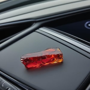 Op de afbeelding: Een ruwe, doorschijnende edelsteen met een vurige rode en oranje tint. Het kristalachtige object ligt op een donker, getextureerd oppervlak, mogelijk in het interieur van een auto. De onregelmatige vorm van de edelsteen vangt het licht.