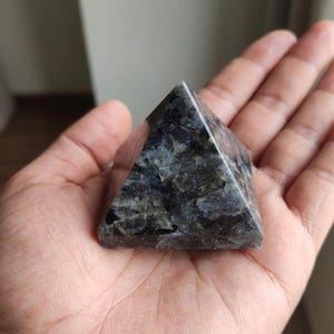 May include: A small, dark stone pyramid held in an open hand. The pyramid has a textured surface with shades of grey, black, and hints of green. The pyramid shape is a classic geometric design, often used in decorative items.