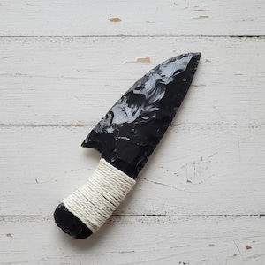 May include: A black obsidian knife with a white wrapped handle. The blade is a dark, glossy black with a sharp point. The handle is wrapped with white twine, providing a secure grip. The knife is displayed on a white wooden surface.