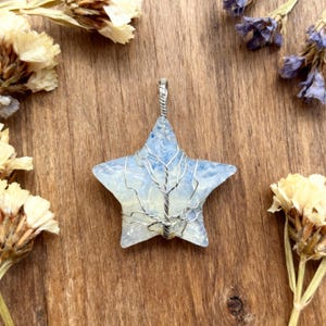 May include: A star-shaped pendant with a light blue and white stone, wrapped in silver-colored wire. The wire is shaped to resemble a tree. The pendant is surrounded by dried flowers on a wooden surface.