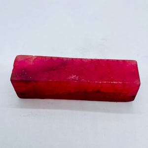 Könnte beinhalten: Ein rechteckiger, roher Rubin-Edelstein mit tiefroter Farbe. Die rauen Kanten und die natürliche Textur des Steins sind sichtbar. Der Rubin wird vor einem weißen Hintergrund präsentiert.
