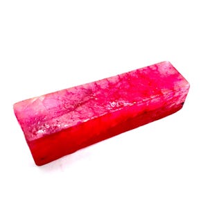 May include: A rectangular, raw ruby gemstone with a vibrant red hue and a slightly translucent appearance. The stone has a rough, natural texture and is set against a white background. The ruby is a gemstone.