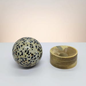 Puede incluir: Una esfera moteada con manchas negras y una base beige claro se encuentra junto a un soporte cilíndrico de madera clara tallada. La esfera es lisa y pulida, mientras que el soporte tiene una textura más rugosa.
