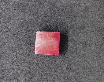 Puede incluir: Un pequeño objeto cuadrado rojo con un acabado brillante. El objeto está sobre un fondo gris oscuro texturizado. El color rojo es vibrante y llama la atención.