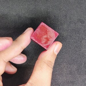 Könnte beinhalten: Ein kleiner, quadratischer, rubinroter Gegenstand wird zwischen zwei Fingern vor einem dunkelgrauen Hintergrund gehalten. Das Objekt scheint fest zu sein, möglicherweise ein Edelstein oder ein dekoratives Material. Die Kanten sind scharf und gut definiert.