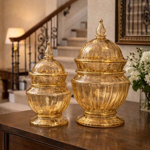 May include: Two decorative glass jars with lids. The jars are amber-colored and sit on a dark wooden table. The jars have a ribbed design and are topped with a pointed finial. The background includes a staircase and a mirror.