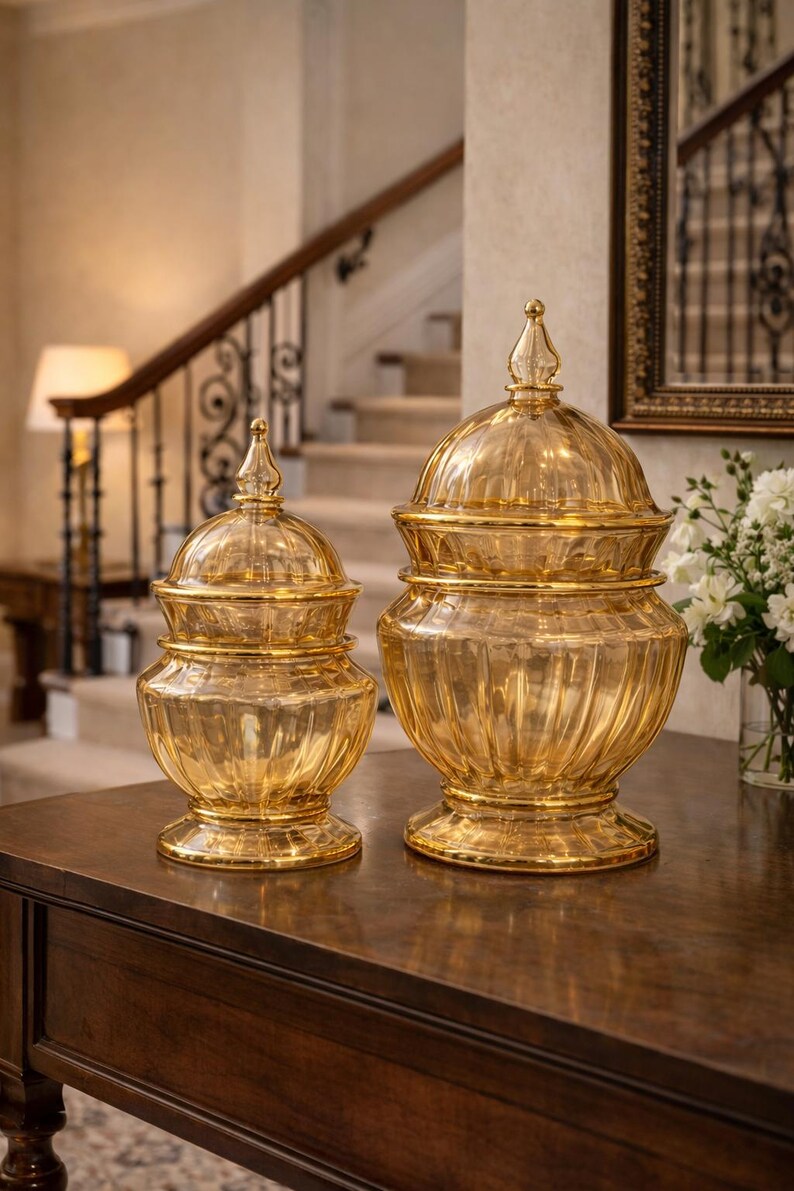 May include: Two decorative glass jars with lids. The jars are amber-colored and sit on a dark wooden table. The jars have a ribbed design and are topped with a pointed finial. The background includes a staircase and a mirror.