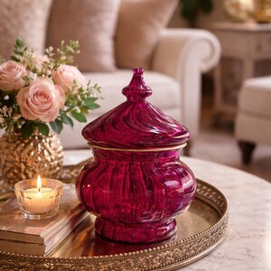 May include: A ruby-coloured glass jar with a lid, displayed on a gold-rimmed tray. The jar features a swirling pattern and gold trim. A lit candle and a bouquet of pink roses in a gold vase are also visible.