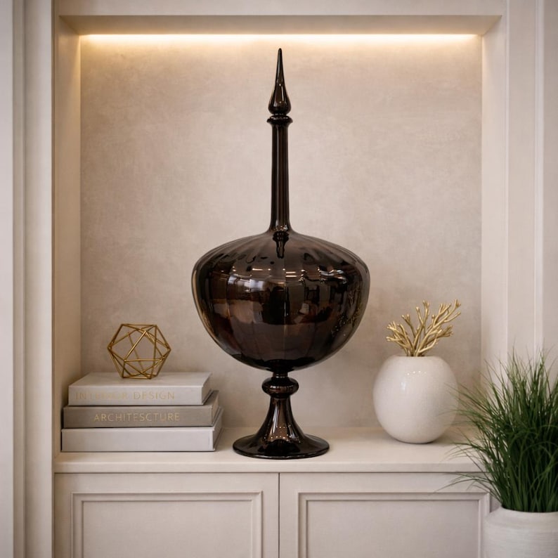 Pu&ograve; includere: Un vaso decorativo alto, di vetro marrone scuro, con un corpo bulboso e un collo lungo e sottile, sormontato da un elemento a punta. Il vaso &egrave; posto su una base. Altri elementi decorativi includono libri, una scultura geometrica dorata e un vaso bianco con rami dorati. Altezza: circa 60 cm.