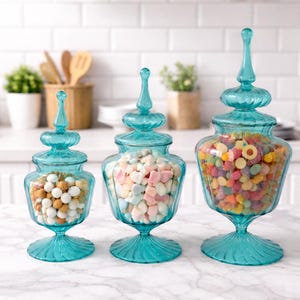 Pode incluir: Três potes de vidro turquesa de tamanhos variados cheios de guloseimas coloridas. Os potes têm tampas ornamentadas e bases de pedestal. Um pote contém doces brancos e bege, outro marshmallows pastéis e o terceiro balas de goma sortidas.