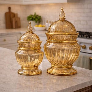 May include: Two decorative glass jars with lids. The jars are amber-colored with gold trim and a ribbed design. The jars are different sizes and sit on a marble countertop in a kitchen setting.