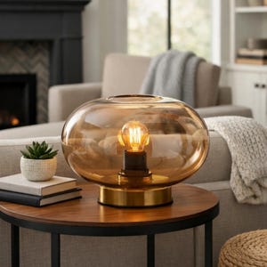 May include: A decorative table lamp with a round, amber-colored glass shade. The lamp features a vintage-style light bulb and a gold-colored base. The lamp is placed on a dark brown, round table.