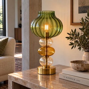 May include: A table lamp featuring a large, ribbed, olive green glass shade atop a brass-finished metal base. The stem consists of three stacked, rounded glass elements in clear and amber tones, illuminated by a single warm-toned light bulb.