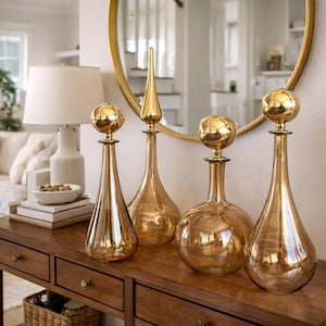 Op de afbeelding: Vier decoratieve glazen flessen met goudkleurige bolvormige stoppen en een gouden afwerking staan op een houten tafel. De flessen variëren in vorm, waarvan één met een puntige top. Een goudkleurige spiegel staat op de achtergrond.