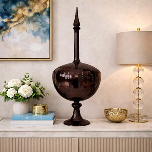 Pu&ograve; includere: Un vaso decorativo marrone scuro con un corpo bulboso e un collo lungo e sottile, sormontato da un pinnacolo appuntito. Il vaso &egrave; esposto su una superficie bianca con altri oggetti decorativi, tra cui una lampada e una composizione floreale.