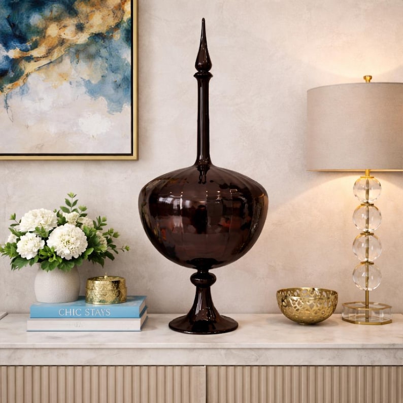 Pu&ograve; includere: Un vaso decorativo marrone scuro con un corpo bulboso e un collo lungo e sottile, sormontato da un pinnacolo appuntito. Il vaso &egrave; esposto su una superficie bianca con altri oggetti decorativi, tra cui una lampada e una composizione floreale.