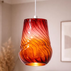 May include: A pendant light with a red and orange glass shade. The shade has a swirling, textured design and a warm glow from the light source. The fixture is suspended from a white cord.