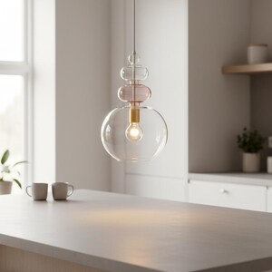 May include: A pendant light fixture featuring a clear glass globe base, a smaller translucent pink glass sphere above it, and a tiny clear glass orb at the top. The light is suspended by a thin cord and includes a brass-toned socket with a glowing bulb.