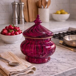 May include: A magenta glass jar with a lid and gold trim, displayed on a marble surface. The jar is part of a kitchen setting, with a spoon, napkin, and a bowl of strawberries nearby. The jar's height is approximately 20 cm.