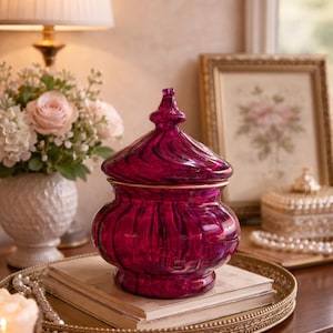 May include: A deep magenta glass jar with a lid, featuring a swirling design and gold trim. The jar sits on a stack of books on a gold-toned tray, alongside a lit candle and a vase of flowers. The jar is a decorative home accent.
