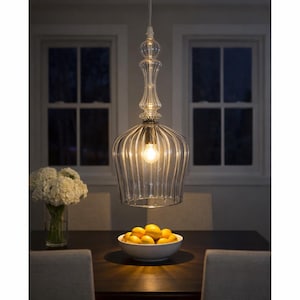 Pode incluir: Uma luminária pendente de vidro transparente com um design decorativo pende sobre uma mesa. A luz ilumina uma tigela de limões e um vaso de flores brancas. Duas janelas são visíveis no fundo.