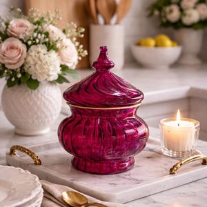 May include: A vibrant, ruby-red glass jar with a lid and gold trim is displayed on a marble tray with gold handles. A lit candle and white plates are also present. The kitchen setting includes flowers and wooden utensils.