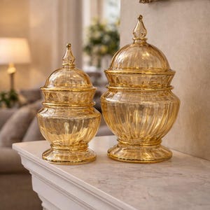 May include: Two decorative glass jars with lids. The jars are amber-colored with a ribbed design and gold accents. They are displayed on a white mantelpiece. The jars have a classic, elegant shape, suitable for home decor.