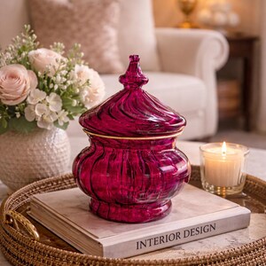 May include: A decorative, ruby-coloured glass jar with a lid, accented with gold trim, sits on a woven tray. The tray also holds a book with "INTERIOR DESIGN" printed on the cover and a lit candle. A vase of flowers is in the background.