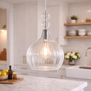 Puede incluir: Lámpara colgante de cristal transparente con diseño acanalado, colgada en una cocina. La lámpara tiene forma de lágrima y una parte superior plateada. La cocina cuenta con armarios blancos, encimera de mármol y una tabla de cortar de madera con limones.