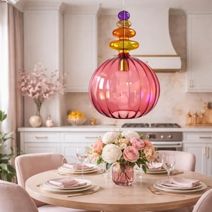 Puede incluir: Una lámpara colgante de cristal rosa con una pantalla esférica acanalada cuelga sobre una mesa de comedor redonda. La lámpara tiene una parte superior apilada y colorida con elementos morados, dorados y naranjas. Un arreglo floral y cubiertos adornan la mesa.