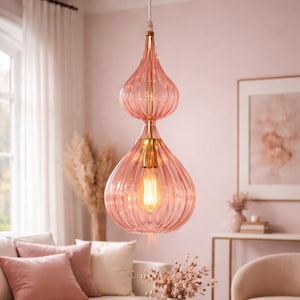Puede incluir: Lámpara colgante de cristal rosa con diseño acanalado y detalles dorados. La lámpara tiene forma de lágrima con una bombilla en el interior, colgando de un cable blanco. La luz está en un salón.