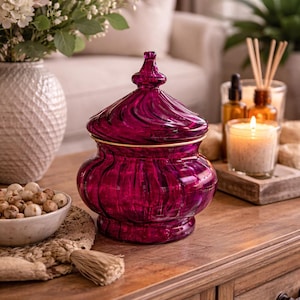 May include: A deep magenta glass jar with a lid and gold trim. The jar has a rounded base and a swirling pattern on the lid. It is on a wooden table, with a bowl of decorative spheres and a lit candle. The jar is likely for decorative purposes.