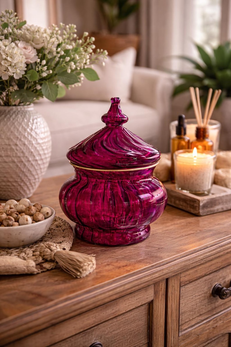 May include: A deep magenta glass jar with a lid and gold trim. The jar has a rounded base and a swirling pattern on the lid. It is on a wooden table, with a bowl of decorative spheres and a lit candle. The jar is likely for decorative purposes.