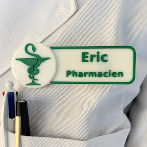 Op de afbeelding: Een wit rechthoekig naamplaatje met een groene rand en een groen caduceussymbool aan de linkerkant. De tekst Eric en Pharmacien is in het groen op het plaatje gedrukt. Het plaatje is bevestigd aan een witte laboratoriumjas met drie pennen.