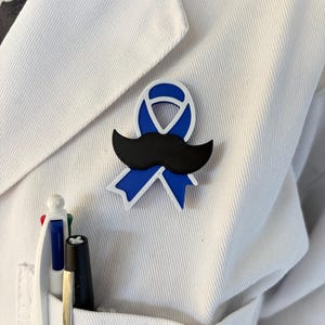 Op de afbeelding: Een blauwe bewustwordingslint-speld met een witte rand en een zwarte snor-opzetstuk, bevestigd aan een wit stoffen kledingstuk. De speld heeft een klassiek lusontwerp met de snor horizontaal geplaatst over het midden van het lint.