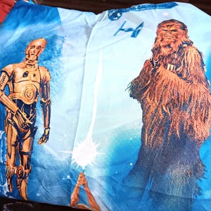 May include: A blue and white Star Wars-themed bed sheet featuring C-3PO and Chewbacca. The sheet has a light blue background with a graphic of a lightsaber and a TIE fighter. C-3PO is gold, and Chewbacca is brown.