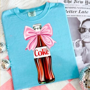 Puede incluir: Una camiseta azul claro con un gráfico de una botella de vidrio de Diet Coke adornada con un gran lazo de satén rosa brillante. La botella tiene el logotipo clásico de Diet Coke en rojo y blanco en la etiqueta. La camiseta tiene una etiqueta Comfort Colors.