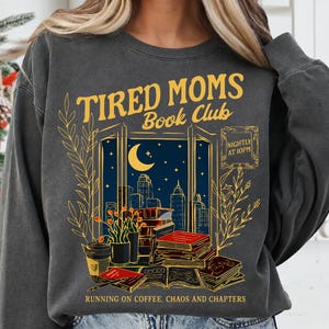 Op de afbeelding: Een antracietgrijze sweater met ronde hals en een grafische print met de tekst TIRED MOMS BOOK CLUB, NIGHTLY AT 10PM en RUNNING ON COFFEE, CHAOS AND CHAPTERS. Het ontwerp toont een stadsgezicht bij nacht, boeken en een koffiebeker.