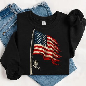 May include: Black sweatshirt featuring a distressed American flag design with a fishing rod graphic. The flag is red, white, and blue, with a detailed illustration of a fishing rod and reel. The sweatshirt is paired with blue denim jeans.