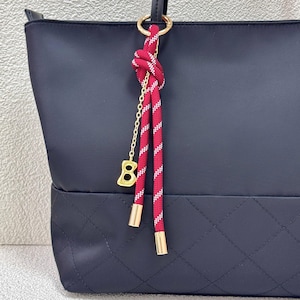 May include: A navy blue tote bag with a quilted bottom panel. A red and white striped rope charm hangs from a gold ring on the handle. The charm has a gold chain and a gold-colored charm shaped like the number eight.