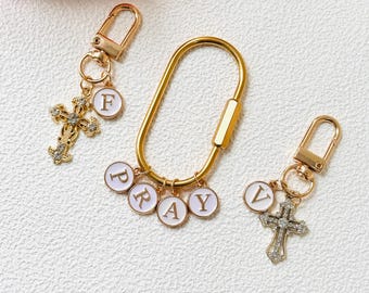 May include: Gold-toned keychains with cross pendants and the word "PRAY". One keychain features a large oval clasp, while the others have smaller clasps. The crosses are embellished with clear crystals and the letters are in a white circle.