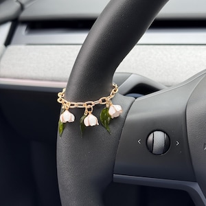 May include: A gold-toned chain bracelet featuring dangling charms shaped like white lily of the valley flowers and dark green leaves. The bracelet is constructed with textured oval links and is displayed wrapped around a dark gray steering wheel.