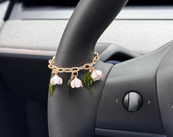 Könnte beinhalten: Ein goldfarbenes Kettenarmband mit baumelnden Anhängern in Form von weißen Maiglöckchenblüten und dunkelgrünen Blättern. Das Armband besteht aus strukturierten ovalen Gliedern und ist um ein dunkelgraues Lenkrad gewickelt dargestellt.