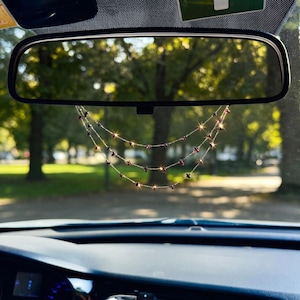 May include: A car rearview mirror with a three-strand beaded car charm. The charm features small red and black beads and is suspended from the mirror. The background shows a blurred view of trees and sunlight.