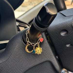 May include: A gold-toned chain charm accessory featuring four dangling pendants: a crescent moon with a face, a teal star, a sun with a face, and a small red heart. The chain is looped around a black plastic vehicle steering column lever.