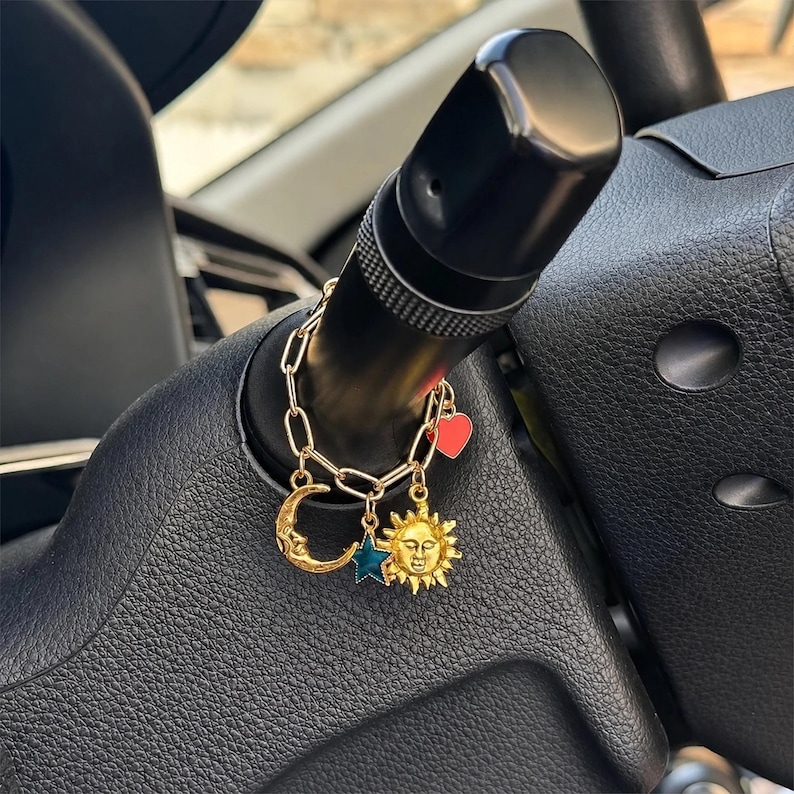 May include: A gold-toned chain charm accessory featuring four dangling pendants: a crescent moon with a face, a teal star, a sun with a face, and a small red heart. The chain is looped around a black plastic vehicle steering column lever.